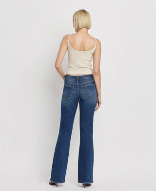 A person with short blonde hair is standing with their back to the camera, wearing a beige sleeveless top, Kip Mid Rise Relaxed Bootcut Jeans by Lovervet by VERVET, and dark shoes. The background is plain white.