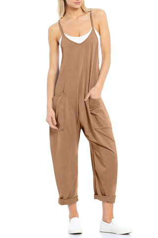 A woman wears a loose, brown Randy Hotshot Onesie V-Neck Romper by Charmo with rolled-up cuffs over a white tank top. She has one hand in a pocket and is wearing white slip-on shoes. The romper features spaghetti straps and large front pockets, perfect for effortless summer looks.