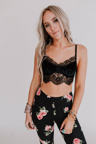 A woman with long blonde hair is wearing the ultra-chic West Velvet and Lace Cami in black by Three Bird Nest, paired with floral-patterned leggings. She accessorizes with several bracelets and rings, striking a confident pose with one hand on her hip against a plain white background, epitomizing the essence of a global fashion oasis.