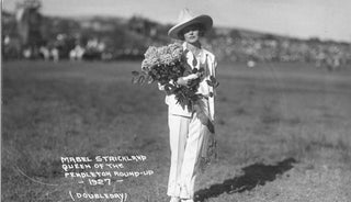 Black and white photo of a woman in a white suit and wide-brimmed hat holding a bouquet of flowers. Text on the image reads, "Inspired by Mabel Strickland, Queen of the Pendleton Round-Up 1927 (Doubleday)." A grassy field and crowd in the United States are visible in the background. Introducing Lady Mabel Perfume by R. Rebellion.