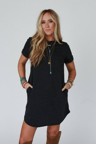 A woman with long blonde hair wearing the "On The Daily Pocket Tee Dress" in charcoal by Three Bird Nest, brown knee-high boots, and multiple colorful bracelets stands against a plain background with her hands in her dress pockets. She is accessorized with layered necklaces and exudes a relaxed silhouette.