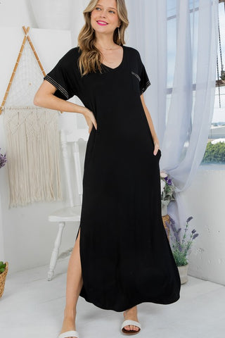 A woman stands indoors in the Faye Curvy Maxi Dress from Western Edge Boutique, a black, short-sleeved, playful maxi dress. She has one hand on her hip and is smiling. The room is adorned with a macramé wall hanging, sheer curtains, and potted plants. Her white slide sandals perfectly complete the comfort of her outfit.