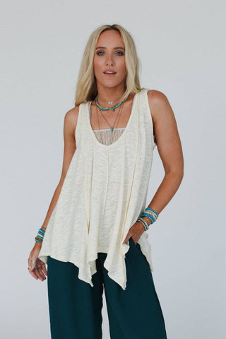 A person with long blonde hair wearing the Three Bird Nest's Elara Top in a neutral cream color and dark green pants, embodying a boho style with layered necklaces and multiple bracelets on both wrists. The background is light grey.