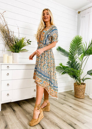 A woman with long blond hair wearing an Anna Kaci Wholesale Finn Tie Back High Low Dress and wedge sandals stands in a stylish room. The walls are white with horizontal slats, and there is a white dresser with potted plants placed on top. A tall, leafy plant in a woven basket stands near her.
