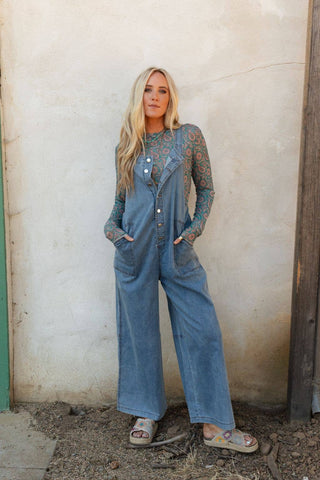 A woman with long blonde hair stands against a textured wall. She wears a sheer, long-sleeved top with a colorful pattern underneath the Dock Town Button Up Jumpsuit - Denim by Three Bird Nest, which features large pockets. With her hands in the pockets and colorful sandals on her feet, she exudes boho style clothing vibes. The ground around her is bare earth.