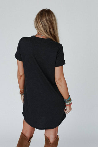 A person stands facing away from the camera, wearing an On The Daily Pocket Tee Dress in charcoal by Three Bird Nest. They have long blonde hair and are accessorized with multiple bracelets on both wrists. The relaxed silhouette is completed by tall brown boots against a plain, light-colored background.