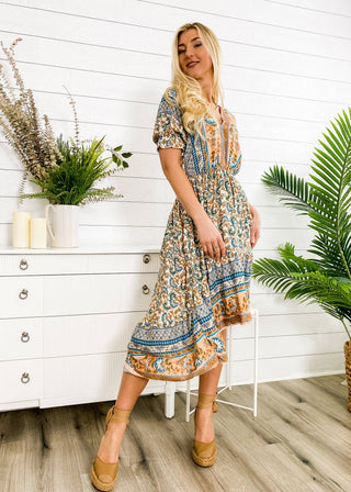 A person with long, blonde hair is wearing the Finn Tie Back High Low Dress by Anna Kaci Wholesale. They are smiling and posed next to a white chest of drawers adorned with plants and candles. The background is a white, shiplap wall enhanced by additional greenery.