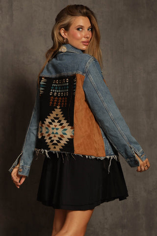 A woman with long hair models the Blue Buttercup Ryder Aztec Print with Faux Suede Denim Jacket, showcasing its colorful geometric back pattern. She pairs it with a black dress and poses against a textured gray background, glancing over her shoulder.