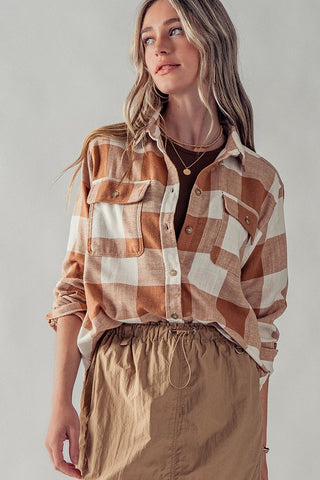 A woman with long blonde hair dons the urban daisy Carmel Checkered Flannel-Fall Flannel over a brown top, paired with a tan skirt. Her autumn look is complemented by the soft, neutral-toned background.