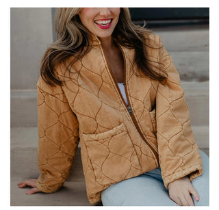 A smiling person with long hair sits on a step, wearing the Kipton Quilted Jacket by Western Edge Boutique in a tan shade, showcasing its visible stitching patterns. The unzipped jacket reveals a white top underneath as their hands rest on their lap.