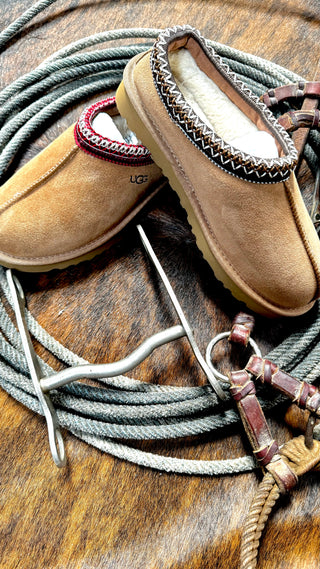 A pair of Bailey Slippers from Western Edge Boutique, featuring a tan slip-on design with patterned trim, rests on a coiled rope. Nearby, leather straps and a metal clip lie on a textured brown surface, creating a rustic atmosphere. These comfortable slippers are the perfect Christmas gift, combining both style and comfort.