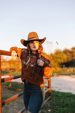 Wearing the Saddle Ranch Sweater by 2FLY and a cowboy hat, a woman leans on a metal fence in a sunlit rural setting. Her long braided hair cascades over her shoulder as she gazes thoughtfully into the distance, surrounded by grass, trees, and a clear sky.