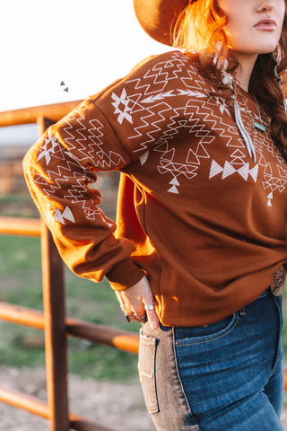 A person wearing the 2FLY Saddle Ranch Sweater, which is brown and adorned with intricate embroidery, paired with blue jeans, stands near a wooden fence. The sunlight highlights their clothes and hair as they wear a hat and earrings.