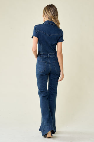 A person with long, wavy hair is standing and facing away from the camera. They are wearing a Women's Virginia Bellbottom Denim Jumpsuit from Western Edge Boutique, featuring a fitted, short-sleeved design with flare legs. The stretchy denim fabric enhances comfort, set against a plain, light-colored background.