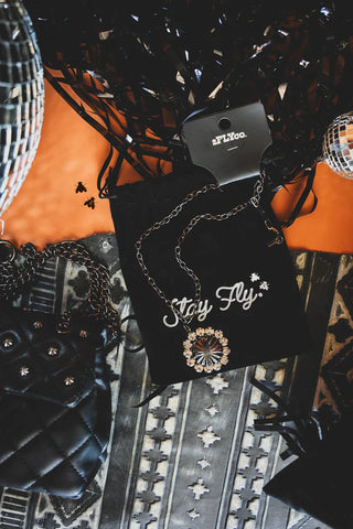 A stylish "Big Bang Theory" necklace with a sparkly pendant is elegantly displayed on a black velvet pouch emblazoned with "Stay Fly." Surrounding the pouch are sleek black and silver accessories, including a quilted handbag, chic earrings, and two disco balls. The setup is set against an intricate patterned fabric backdrop, showcasing the brand 2 Fly’s exquisite taste.