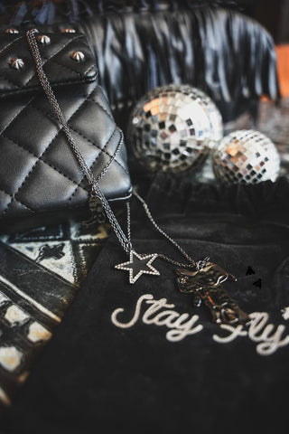 A quilted black handbag with studded details, BLAZIN' SADDLE NECKLACES by 2 Fly featuring silver star and cross designs, and two mirrored disco balls are arranged on a fabric background. The words "Stay Fly" are stitched onto a piece of fabric beneath the quality jewelry, perfect for those who love to mix and match.