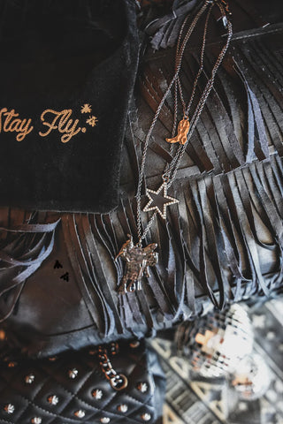 Image features a black velvet pouch embroidered with "Stay Fly," alongside several quality 2 Fly BLAZIN' SADDLE NECKLACES adorned with charms including an airplane, a star, and a bird, all placed on a fringed black leather surface. A studded black accessory and a small disco ball are partially visible, perfect for mixing and matching.