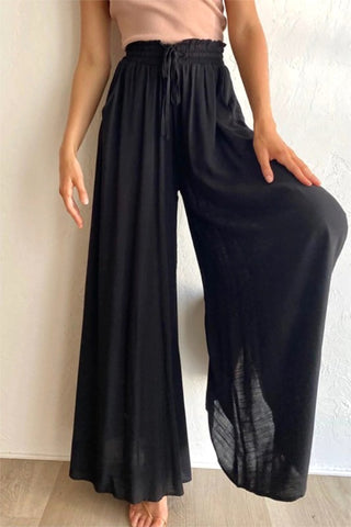 A person wearing the TFY Collection's black Prairie Wide Leg Palazzo Pants, paired with a light pink top, is posing against a plain white wall. The flowy pants, which can appear slightly see-through under bright light, feature a drawstring waistband for maximum comfort. Their left hand rests on their hip.