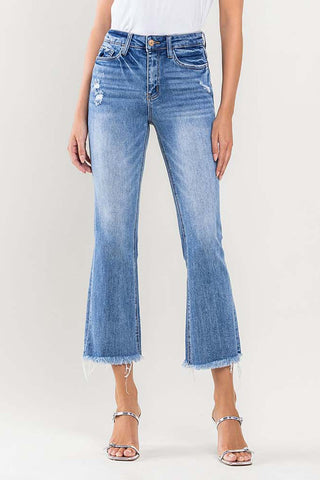 A person is dressed in a white top paired with the Nash High Rise Stretch Curvy jeans from Western Edge Boutique. These blue, high-rise, distressed kick flare jeans feature a frayed hem along with subtle rips and a faded look. The ensemble is completed with silver strappy high-heeled sandals, set against a plain background.