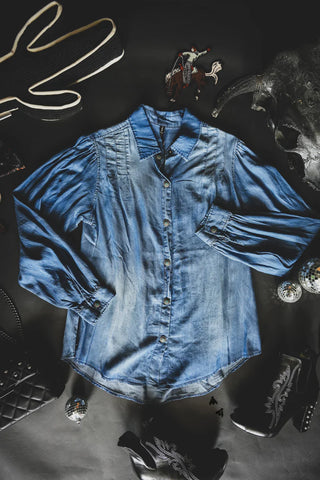 A neatly spread COWBOY BLUES chambray button-up shirt by 2 Fly with long sleeves is placed on a dark surface. Surrounding it are various items, including a cowboy hat, ankle boots with Western designs, a quilted black handbag, small decorative items, and a cactus-shaped lamp.