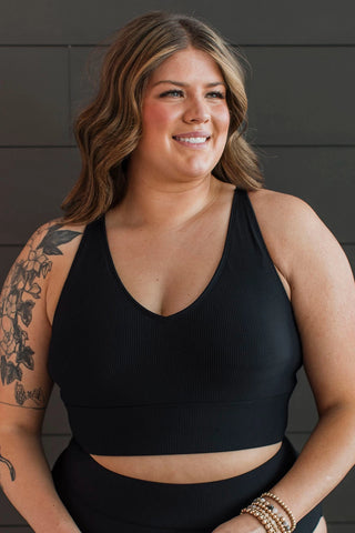 A smiling woman with wavy hair is wearing the Kissed By The Sun Bikini Swim Top in black ribbed from Western Edge Boutique. She stands against a dark wall, showcasing her floral tattoo on her arm and multiple bracelets.