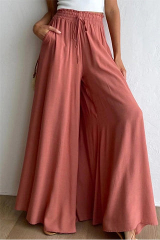 A person is wearing TFY Collection's Prairie Wide Leg Palazzo Pants in rust. These high-waisted, prairie-inspired pants feature a wide-leg design, an elastic waistband with a drawstring, and are complemented by a tucked-in white top. The person's hands rest comfortably in the pockets of the pants.