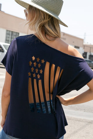 A woman stands outdoors wearing a wide-brimmed hat and Western Edge Boutique's Evie Curvy 4th of July Laser Cut-Out Top in navy blue. The loose-fit top drapes effortlessly as she faces away from the camera, showcasing its patriotic American flag design on the back, one hand resting confidently on her hip.