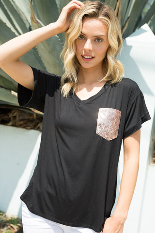 A woman with blonde hair stands outdoors, wearing the Lyndey Curvy Black Sequin Pocket Boxy Top from Western Edge Boutique paired with white pants. She is touching her hair with her right hand and looking at the camera. Large, green leaves are visible in the background.