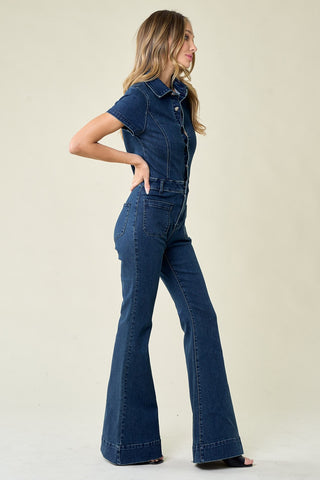 A woman stands in profile, donning a Women's Virginia Bellbottom Denim Jumpsuit by Western Edge Boutique. Her long, wavy blonde hair cascades as she poses with one hand on her hip, looking into the distance. The short sleeves and flared legs highlight her chic style, while the stretchy denim fabric ensures comfort against the plain, light-colored background.