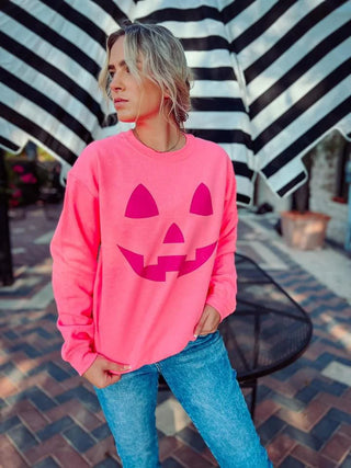 A person stands confidently outdoors wearing the "Neon Jack Hot off the Press" sweatshirt from Western Edge Boutique, featuring a neon pink design with a purple jack-o'-lantern face on the front. Paired with blue jeans, they stand before a large black and white striped umbrella, perfectly embodying the Halloween spirit.