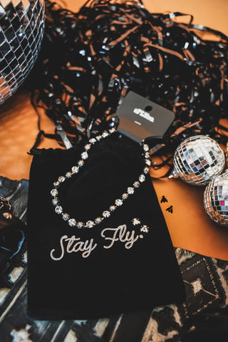 A Dolly Shines Necklace - Western concho sparkle from 2 Fly is elegantly displayed atop a black velvet pouch adorned with the words "Stay Fly" in sparkling letters. Nearby, two small disco balls and a tangled string of black ribbon add flair, all set against an edgy orange background. The scene exudes jewelzzz with attitude.