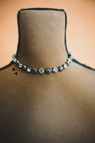 The Dolly Shines Necklace - Western concho sparkle by 2 Fly is displayed on a brown mannequin, featuring alternating clear and dark beads. This choker-style necklace stands out against the plain background, adding an edgy flair to the sparkling jewels.