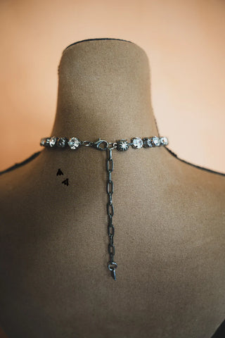 A close-up of a mannequin displaying an edgy, sparkling Dolly Shines Necklace - Western concho sparkle by 2 Fly. The choker, adorned with large, round, crystal-like gems and a chain extension hanging down the back, exudes sophistication. The background is a soft, warm hue, highlighting the elegance of this piece from jewelzzz.