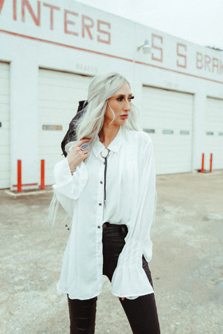 A woman with long blonde hair stands outdoors, wearing the WYOMING DAZE White Button Up Top by 2FLY and black pants. She is holding the strap of a black bag that is slung over her shoulder. Behind her is a white building with garage doors and faded red lettering at the top.