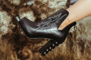 A person's leg wearing the Western Edge Boutique's HIGH DEMAND *LUX black leather ankle boot is extended towards the camera. The high-quality boot boasts intricate white stitching and metal studs on the chunky heel. A small triangle tattoo adorns the person's ankle, while a cactus can be seen in the blurry background.