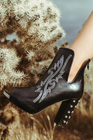 The HIGH DEMAND *LUX black leather ankle boot from Western Edge Boutique is showcased against a background of desert flora, including a blurred cactus. This high-quality ankle boot is adorned with intricate white embroidery and decorative metal studs on the heel. Featuring a high, chunky peep toe heel, it exudes edgy elegance.