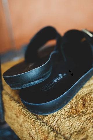 A close-up of the Slick Lux Black Slides by Western Edge Boutique resting on a cushion, showcasing their western buckles, sophisticated sole, and chic straps. The softly blurred background enhances the warm and relaxed atmosphere, creating a truly chef's kiss moment.