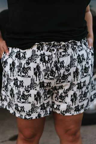 A person is shown from the waist down, wearing Western Edge Boutique's "Gone With The Winds" white paperbag shorts adorned with a black cowboy and desert print, paired with a black top. The shorts feature a COMFYYY stretchy waistband, with hands placed on the sides.