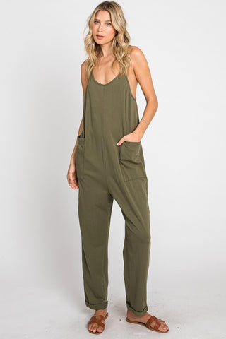 A woman with long blonde hair stands posing in a studio. She is wearing a dark olive green Ledger Harlem Cami Relaxed Jumpsuit by Finaltouch Apparel, featuring two front pockets and brown sandals. The jumpsuit has thin straps and a relaxed fit. The background is white.