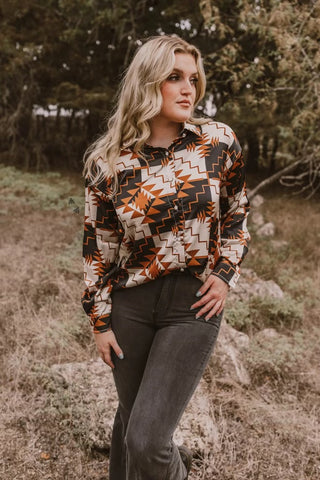 A woman with long, wavy blonde hair stands outdoors in front of trees, wearing the 2 Fly Prexton Aztec Button silky long sleeve top featuring a southwestern pattern in shades of orange, brown, and cream. She pairs it with dark pants, rests one hand on her thigh, and gazes off to the side, exuding western vibes.