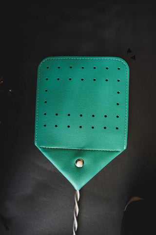 A Western Boot Stitch FLY SWAT IT fly swatter by 2 Fly, made from green cowhide leather with multiple small round holes, a central metal button, and a twisted handle is positioned vertically against a dark background. Several small black specks, possibly insects, are near the top right corner of the image.