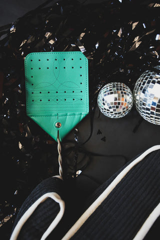 A Western Boot Stitch FLY SWAT IT by 2 Fly is positioned on a black surface with a cowhide-patterned item in the foreground. Two small disco balls are to the right of the fly swatter, while crumpled black plastic sheets or streamers surround the items.