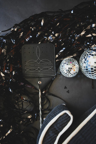 A close-up photo showcases the 2 Fly Western Boot Stitch FLY SWAT IT, a black leather flyswatter crafted from cowhide with white stitching, lying on a bed of black tinsel. Nearby are two small disco balls, tiny silhouettes of people, and a rolled-up black and white-striped fabric. The dark, slightly reflective background highlights the product sharply.