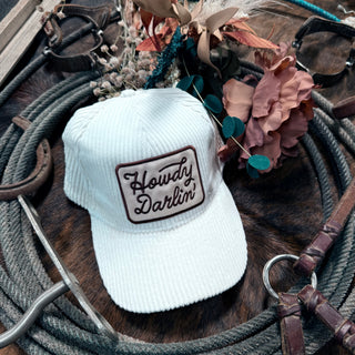 A Howdy Darlin hat by Western Edge Boutique, featuring a white corduroy design with "Howdy Darlin'" text, is artfully placed on a coiled rope and accompanied by dried flowers and leather straps on a wooden surface, creating a rustic accessory statement.