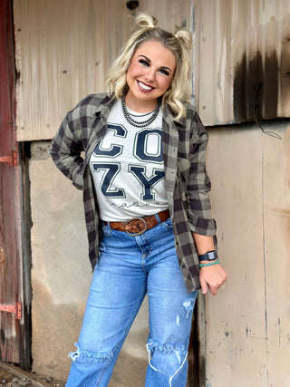 A blonde woman with two ponytails smiles at the camera. She wears an oversized black and white checkered flannel shirt from Western Edge Boutique's Everyday Comfort Flannel collection over a "COZY" t-shirt, a brown belt, distressed blue jeans, and an Apple Watch. The rustic background features a weathered wall with peeling paint, enhancing her stylish outfit.