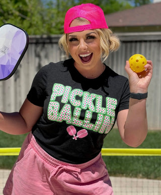 A person in a pink cap and a Western Edge Boutique "Pickle Ballin'" shirt beams with joy while holding a pickleball paddle and ball. They are positioned in front of the pickleball net, with a wooden fence and vibrant green grass setting the ideal scene.