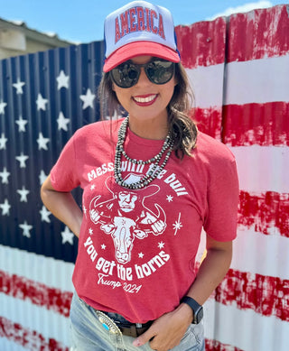 A woman stands in front of a painted American flag backdrop, wearing a red T-shirt from Western Edge Boutique that speaks for itself with a bull illustration and the text "Mess With The Bull You Get The Horns." She's also sporting a red and white "America" cap, sunglasses, and a beaded necklace. She is smiling.