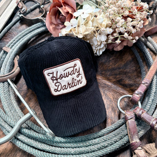 A corduroy hat from Western Edge Boutique, featuring the text "Howdy Darlin," rests atop a coil of rope. Dried flowers, a leather belt, and metal tools are arranged on a textured surface, enhancing the rustic western vibe of this charming accessory scene.