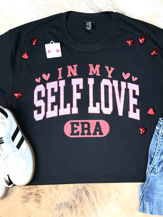 The "In My Self Love Era" black unisex tee from Western Edge Boutique features pink and red text with small red hearts. It's styled on a soft beige surface alongside white sneakers and blue jeans, embodying self-rediscovery.