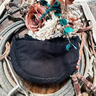 The Corduroy Cross Body from Western Edge Boutique is elegantly displayed on a coiled rope, surrounded by brown leather straps and accented with a rustic floral arrangement of pink and white flowers amidst green leaves on the wooden surface. The scene bursts with multiple colors, creating a vivid and inviting tableau.